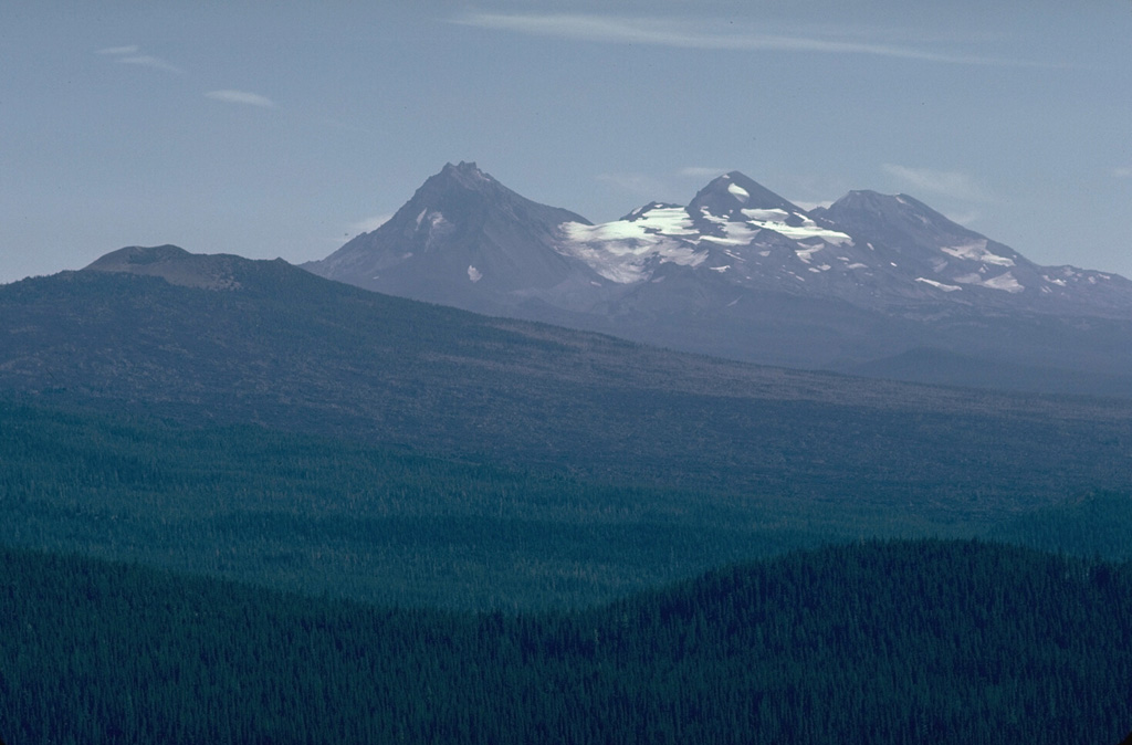 The broad Belknap shield volcano to the left seen here from Sand Mountain volcano to its NW, with the Three Sisters volcanoes in the background, is one of the youngest volcanoes in the central Oregon Cascades. The latest dated lava flow traveled 15 km W to the McKenzie River from a vent on the NE side of Belknap about 1,500 years ago. Photo by Lee Siebert, 1981 (Smithsonian Institution).