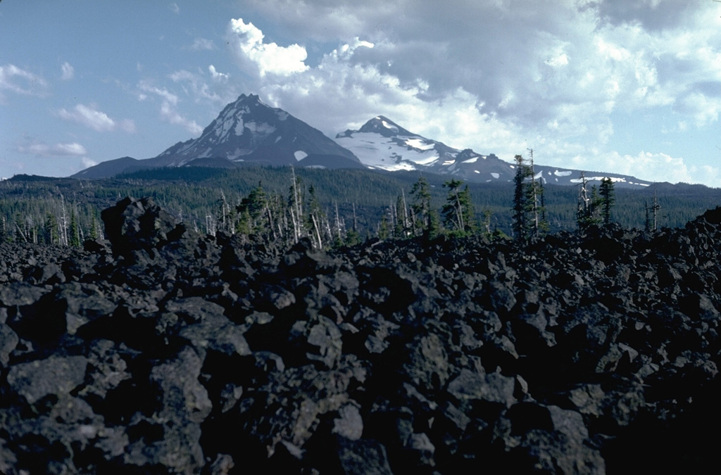 The McKenzie Pass area contains one of the largest concentrations of recent volcanism in the continental United States. Fresh lava flows in the foreground from the Belknap shield volcano merge with unvegetated lava flows from scoria cones on the north flank of North Sister in the background, with Middle Sister to its right. Photo by Lee Siebert, 1982 (Smithsonian Institution).