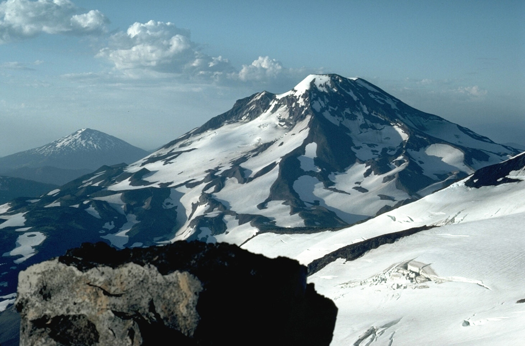 The close proximity of the Three Sisters volcanoes, in contrast to the other large Cascade volcanoes that are typically 60-100 km apart, can be seen in this view of South Sister volcano from the summit of North Sister with the glacier-covered slopes of Middle Sister to the lower right. Mount Bachelor is in the distance to the left. Photo by Lee Siebert, 1984 (Smithsonian Institution).