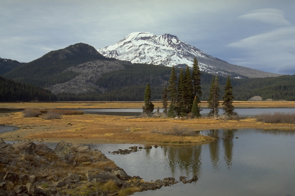The southernmost of a series of obsidian lava domes and flows erupted about 2,000 years ago along a 5-km-long N-S fissure on the SE flank of South Sister, with the end of the flow visible to the left near Sparks Lake. The barren lower right skyline of South Sister is the Newberry lava flow, the largest and northernmost of the lava flows erupted along the 5-km-long chain named after Devils Hill, the forested peak to the left. Photo by Lee Siebert, 1995 (Smithsonian Institution).