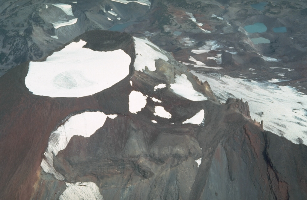 An aerial view of the summit of South Sister from the south, seen here in 1981, shows a snow-filled summit crater that seasonally contains a small melt-water lake. Fissure eruptions occurred during the Holocene from flank vents including some near Carver Lakes on the NE flank, seen here to the upper right. Photo by Willie Scott, 1981 (U.S. Geological Survey).