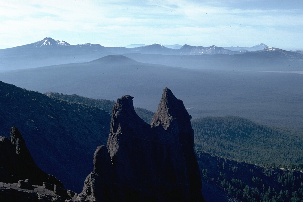 The rim of the Crater Lake caldera is seen in the distance from the summit of Mt. Thielsen to the north. The caldera formed about 7,700 years ago during a large explosive eruption that resulted in the collapse of ancestral Mount Mazama. Mount Scott forms the high point on the left skyline, and Timber Crater is the smaller cone below and left of the north caldera rim. Photo by Lee Siebert, 1982 (Smithsonian Institution).
