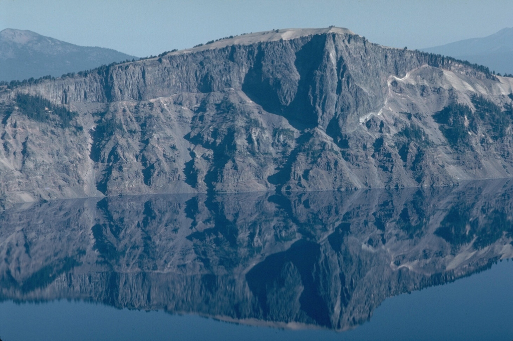 The massive Llao Rock lava flow, exposed in the NW wall of Crater Lake caldera, was emplaced at the end of a major eruption about 200 years prior to the formation of the caldera. The thin, light-colored unit at the base of the lava flow, seen prominently on the right, is a Plinian pumice deposit from that major explosive eruption. The lava flow is more than 350 m thick and is overlain by tephra from the caldera-forming eruption of Crater Lake. Photo by Lee Siebert, 1981 (Smithsonian Institution).