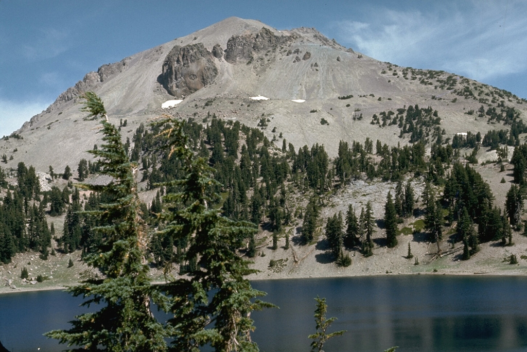 The massive dacitic lava dome of Lassen Peak rises above Lake Helen on the south side of the volcano.  Plugs of dark dacitic lava exposed near the summit are surrounded by vast aprons of lighter-colored talus associated with growth of the lava dome. Photo by Lee Siebert, 1972 (Smithsonian Institution).