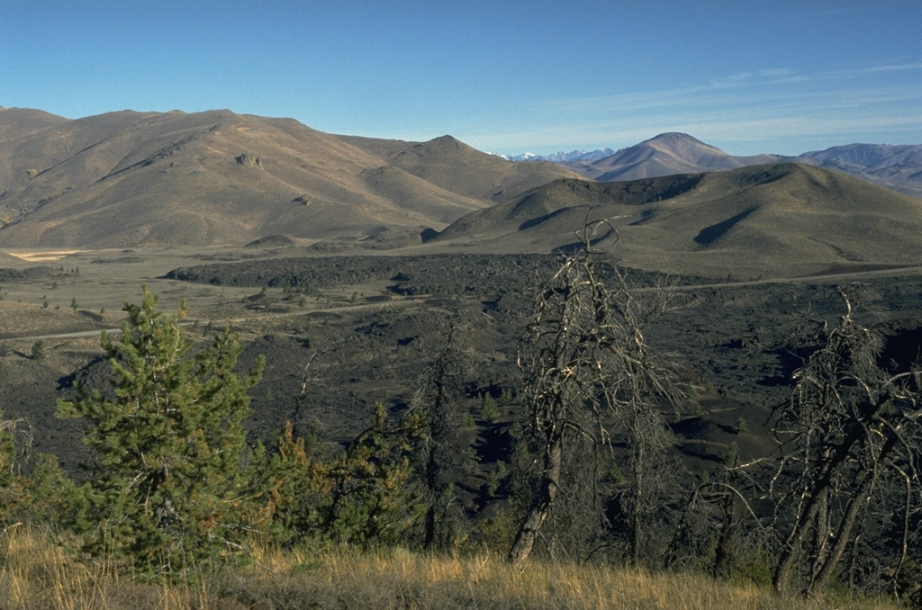 The Highway lava flow forming the black lobe in the center of the photo at the base of the Pioneer Mountains was erupted about 2300 years ago from a vent at or near North Crater, out of view to the right.  Two other Craters of the Moon flows, the Devils Orchard and Serrate lava flows, were also erupted at about the same time.  The complex vegetated cinder cone to the right of the Highway lava flow is the late Pleistocene Sunset Cone. Photo by Lee Siebert, 1994 (Smithsonian Institution).