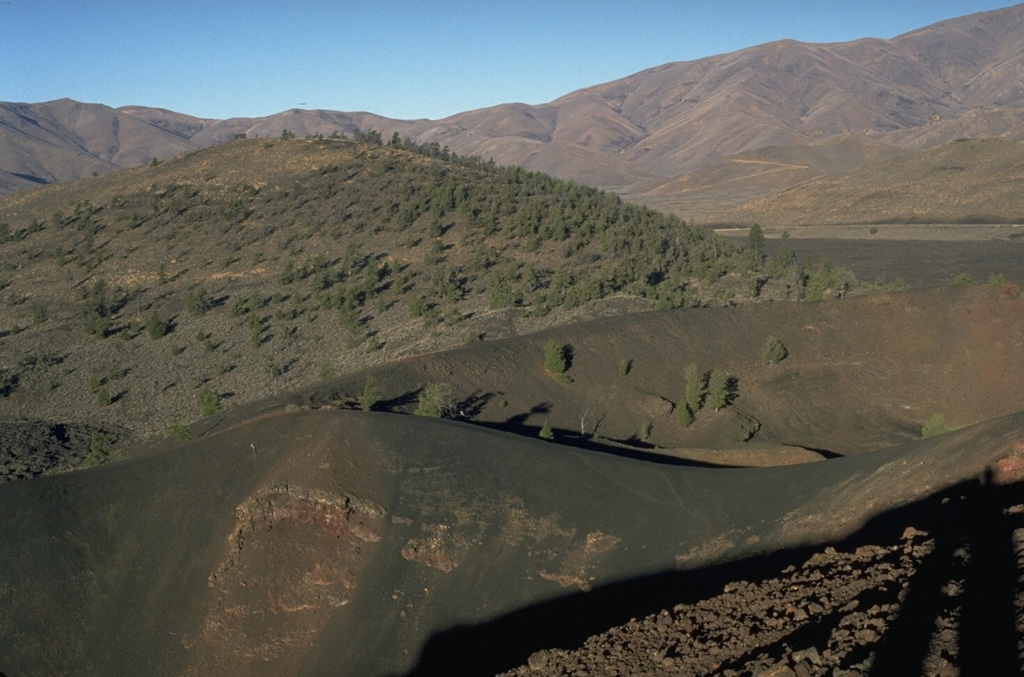 Silent Cone, the partially forested cinder cone in the background, was the source of lava flows primarily to the south about 6500 years ago.  The well-preserved cone of Big Craters in the foreground is one of the youngest features of Craters of the Moon. Photo by Lee Siebert, 1994 (Smithsonian Institution).