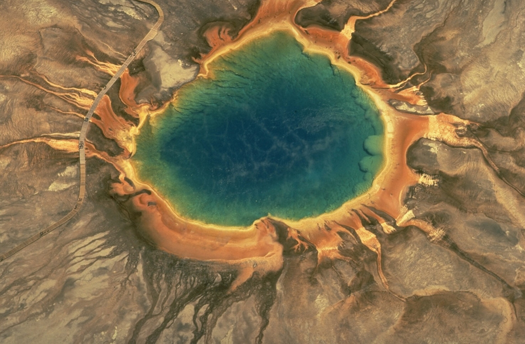 Grand Prismatic Spring, the largest of the geothermal pools in Yellowstone National Park, is 90 m across.  The hot pool lies at the top of a broad, low mound formed by deposition of silica at its margins.  Overflow channels radiate away from the pool.  The colorful rings around the hot-spring pool are formed by algae and other organisms that are temperature dependent.  The highest temperature organisms (70-73 degrees Centigrade) are white.  Lower-temperature organisms grade to light yellow, orange, red, and then dark green. Copyrighted photo by Katia and Maurice Krafft, 1984.