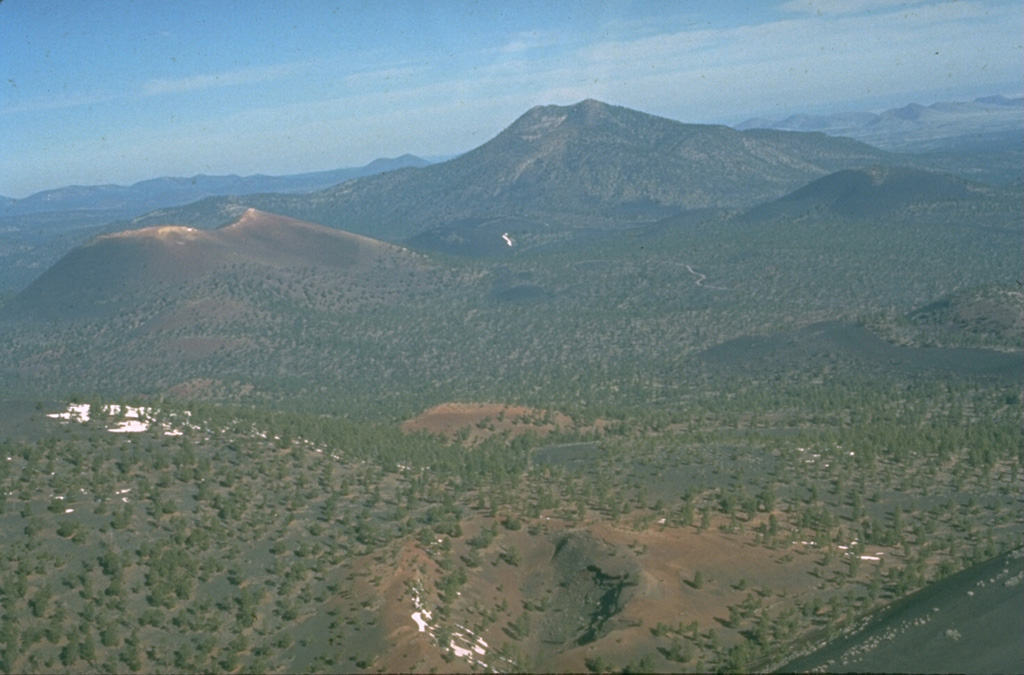 The 60-m-deep Gyp Crater in the foreground is located at about the midpoint of a 10-km-long eruptive fissure extending SE from Sunset Crater, the unvegetated cinder cone at the left-center.  Paleomagnetic evidence places the age of the Sunset Crater eruption at between about 1080 and 1150 CE.  O'Leary Peak, a Pleistocene lava dome, forms the prominent peak in the right background. Photo by Ed Wolfe, 1973 (U.S. Geological Survey).