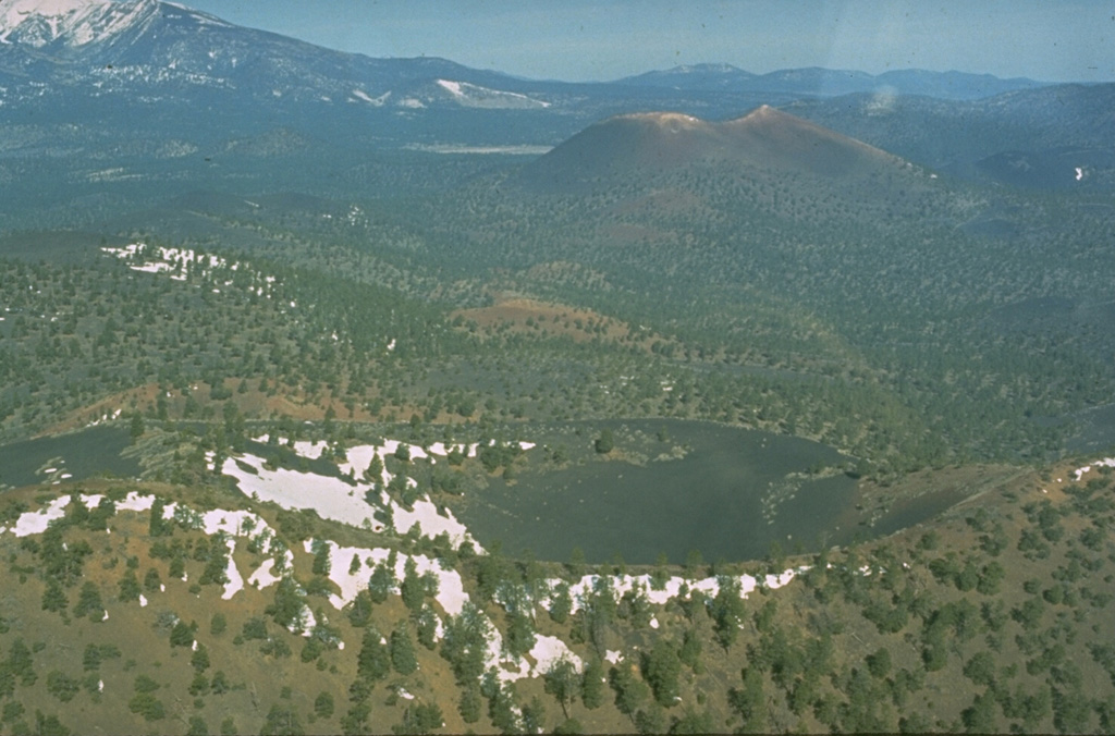 Sunset Crater at the upper right is seen in this aerial view along a 10-km-long eruptive fissure extending to its SE.  An eruption from Gyp Crater, immediately behind the left rim of the snow-dappled Pleistocene Double Crater in the foreground, occurred along the SE-trending fissure.  Snow-capped San Francisco Mountain appears in the background at the upper left. Photo by Ed Wolfe, 1973 (U.S. Geological Survey).