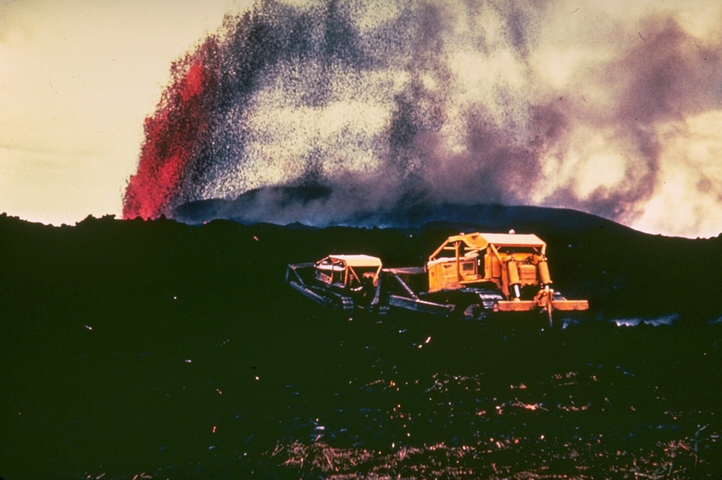 A bulldozer works to create a barrier to lava flows threatening areas below the village of Kapoho, on the lower East Rift Zone of Kilauea, in January 1960. Lava fountains tower in the background from the eruptive vent, which was less than 1 km from the center of the village. Lava flows eventually engulfed the entire village, but the 5 km of walls that were constructed may have helped prevent the flows from reaching houses and a lighthouse at the coast. Photo by Hawaii Volcanoes National Park, 1960.