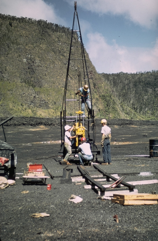 A Hawaiian Volcano Observatory team uses a drilling rig to extract drill core from the cooling lava lake in Kilauea Iki crater. At the time of this 1968 project, nearly a decade after a lava lake filled Kilauea Iki during the 1959 eruption, the crust had solidified to a depth of about 30 m. The drill core penetrated to 60 m depth without reaching the bottom of the still partially molten lava lake. This project, the first to use a drill rig to sample a lava lake, allowed study of vertical variations in chemistry, mineralogy, and temperature within a cooling lava lake. Photo by Jean Tobin, 1968.