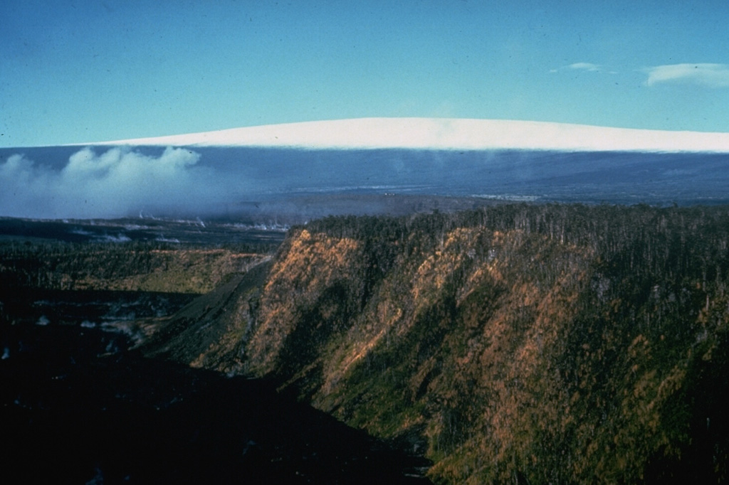Kilauea Iki crater in the foreground was the site of a major eruption in 1959. The crater is in Kilauea's East Rift Zone and is seen here from the east with snow-capped Mauna Loa in the background. Gases rise from the Kilauea caldera to the left.  Photo by Richard Fiske, 1967 (Smithsonian Institution).
