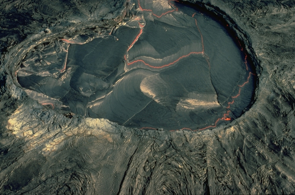 The Kupaianaha lava pond was active for many years during the east rift zone eruption of Kilauea, which began in 1983.  This February 1987 aerial view shows the crusted-over surface of the pond, with incandescent lava visible between segments of the crust.  Convection of the lava lake produces lateral movement and descent of the solidified crust, which mimic many plate tectonic processes.  The smooth slopes surrounding Kupaianaha were formed by overflows of pahoehoe lava. Copyrighted photo by Katia and Maurice Krafft, 1987.