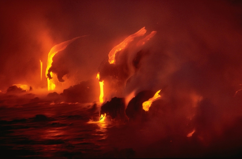 Lava flows dripping over low coastal cliffs are reflected in the sea in this May 1987 nighttime photo.  Flows from Kilauea's east rift zone have repeatedly reached the sea over a broad front during an eruption that began in 1983. Copyrighted photo by Katia and Maurice Krafft, 1987.
