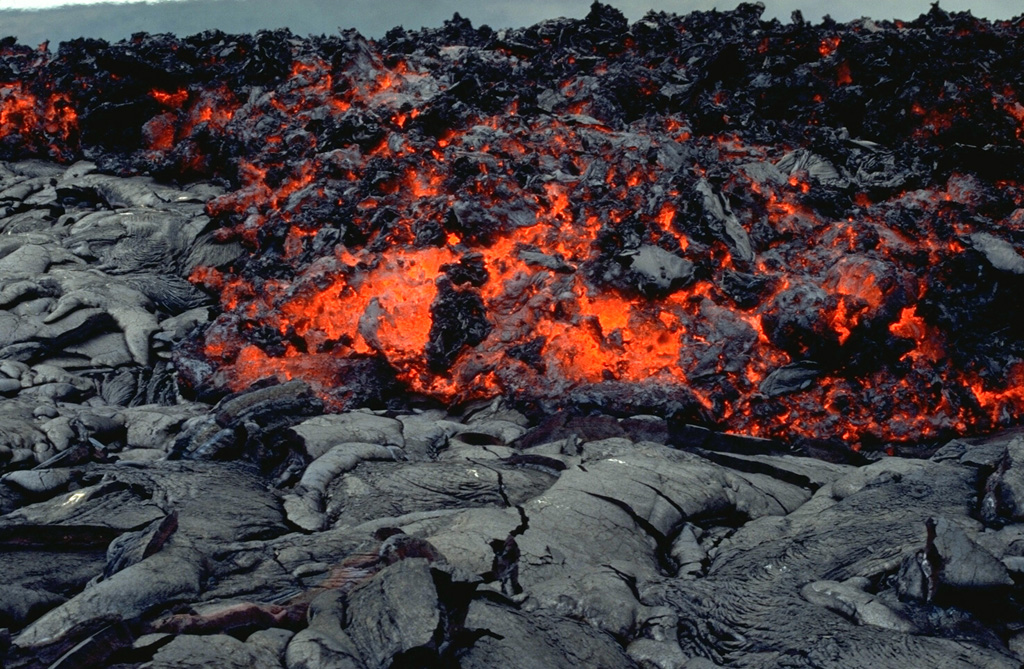 An `a`a lava flow, with a characteristic surface containing abundant angular chunks as it cools, advancing across a smooth-textured pahoehoe lava flow. The hot, incandescent flow interior is visible. The front of this 3 June 1994 flow at Laeapuki, near the Puna coast of Kilauea volcano, is about 1 m thick.  Photo by Paul Kimberly, 1994 (Smithsonian Institution).