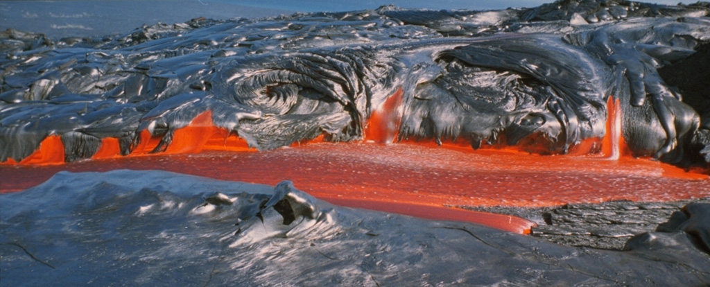 The black, newly solidified glassy surface of pahoehoe lava flows commonly has a silvery or iridescent color. Pahoehoe lavas form during eruptions that are characterized by the upwelling of hot, low-viscosity magma. This smooth-textured pahoehoe flow at Kilauea volcano was photographed in August 1994.   Photo by Paul Kimberly, 1994 (Smithsonian Institution).