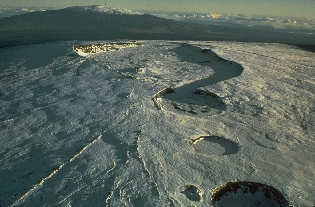 This 1975 photo of the snow-covered summit of Mauna Loa from the SW shows the 2.4 x 4.8 km wide Moku‘āweoweo caldera in the center, with three circular craters in the foreground. South Pit (merging with the SW caldera rim), Lua Hohonu, and Hua Hou (foreground), formed through collapse along the upper SW rift zone. Mauna Kea is in the distance. Photo by Don Peterson (U.S. Geological Survey).