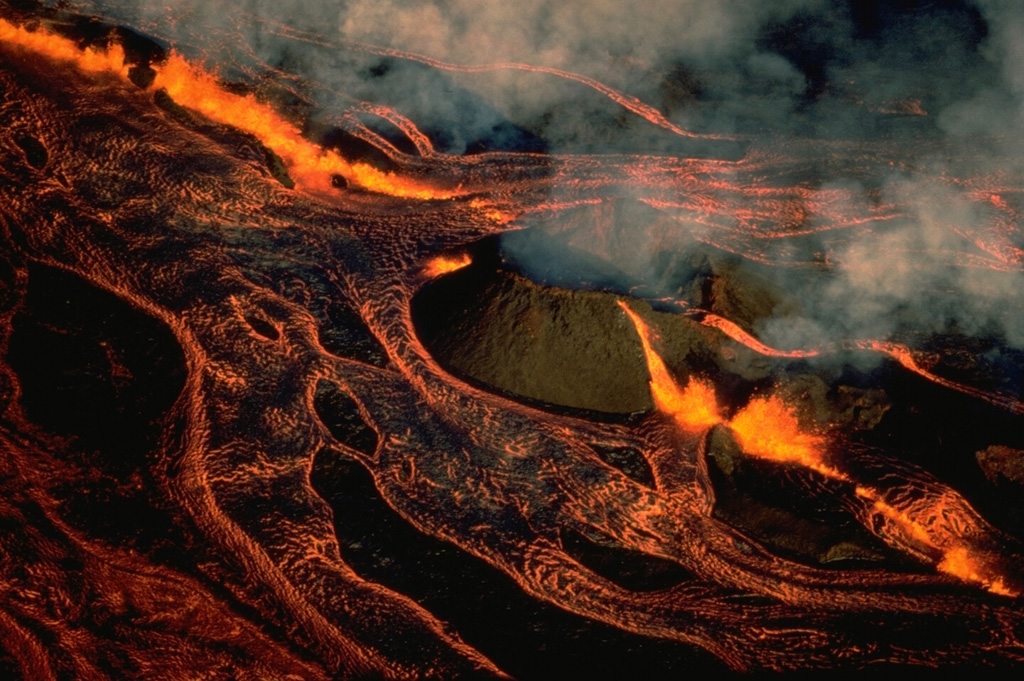 An en-echelon fissure cuts through Pohaku Hanalei, a pre-existing cinder cone on the NE rift zone of Mauna Loa volcano, at dawn on March 25, 1984, during the opening hours of the 1984 eruption.  A line of lava fountains feeds incandescent lava flows that are diverted around the old cone and cover almost the entire field of view in this west-looking photo.  A 2-km-long curtain of fire between about 3400 and 3470 m altitude was active on the morning of March 25, producing lava fountains 10-50 m high. Copyrighted photo by Katia and Maurice Krafft, 1984.