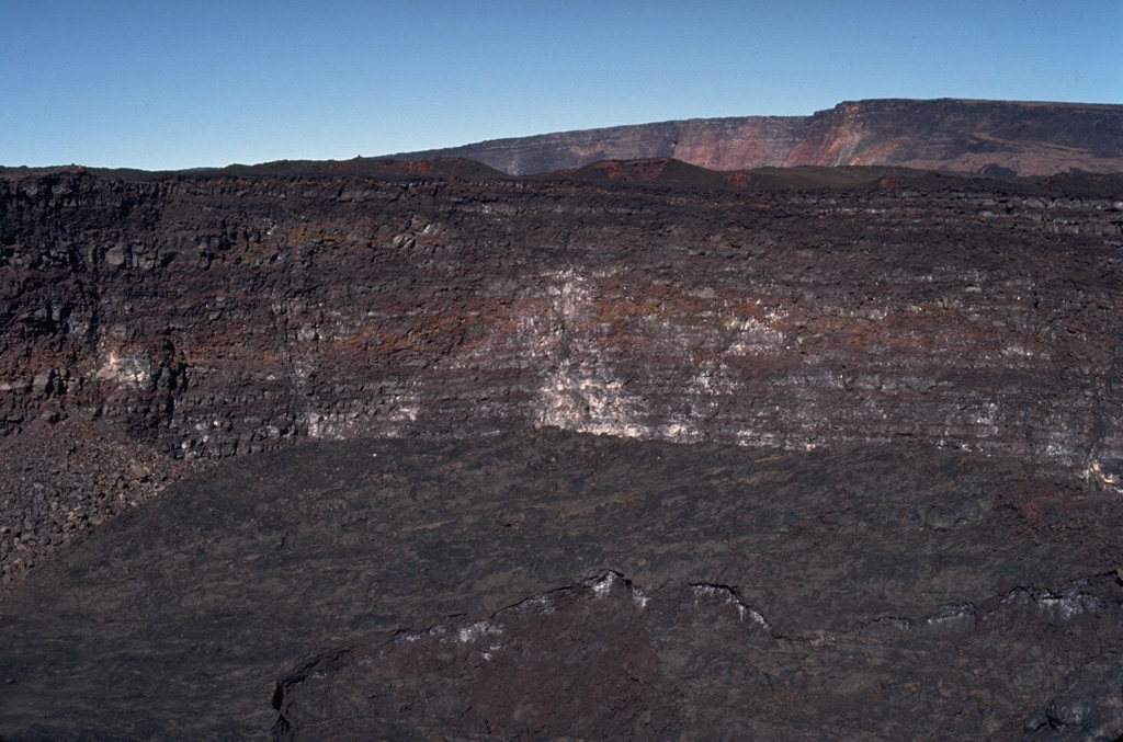 The steep walls of Lua Poholo pit crater, immediately NE of Mokuʻāweoweo caldera, expose a small portion of the accumulation of thin, overlapping lava flows that form Mauna Loa. This view from the NE shows the caldera rim to the upper right. Lava flows from recent eruptions fill crater floor, including the most recent 1984 eruption. Photo by Paul Kimberly, 1994  (Smithsonian Institution).