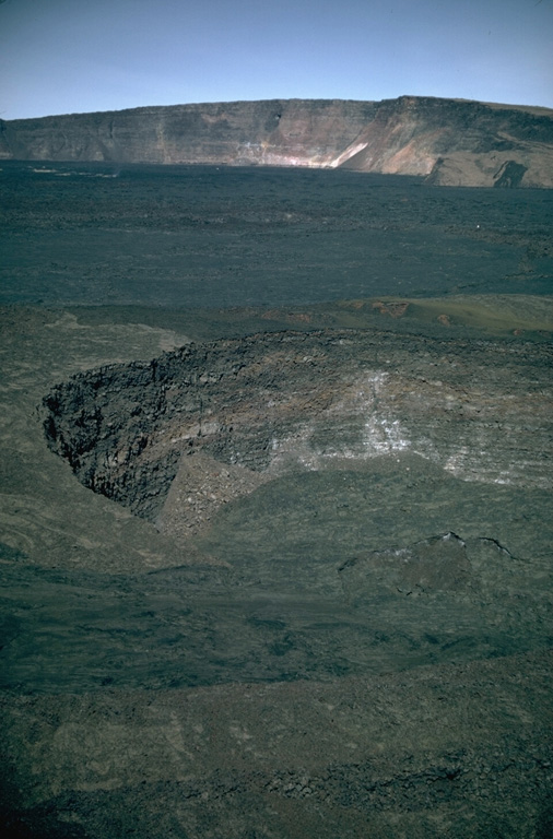 Lua Poholo pit crater is immediately NE of Moku’aweoweo caldera. The crater walls expose a small portion of the massive pile of thin, overlapping lava flows that have formed the summit of Mauna Loa. This 1987 view from the NE shows the western Moku’aweoweo caldera rim. The lava flows covering the caldera floor of the caldera were erupted in 1984. Photo by Richard Fiske, 1987 (Smithsonian Institution).