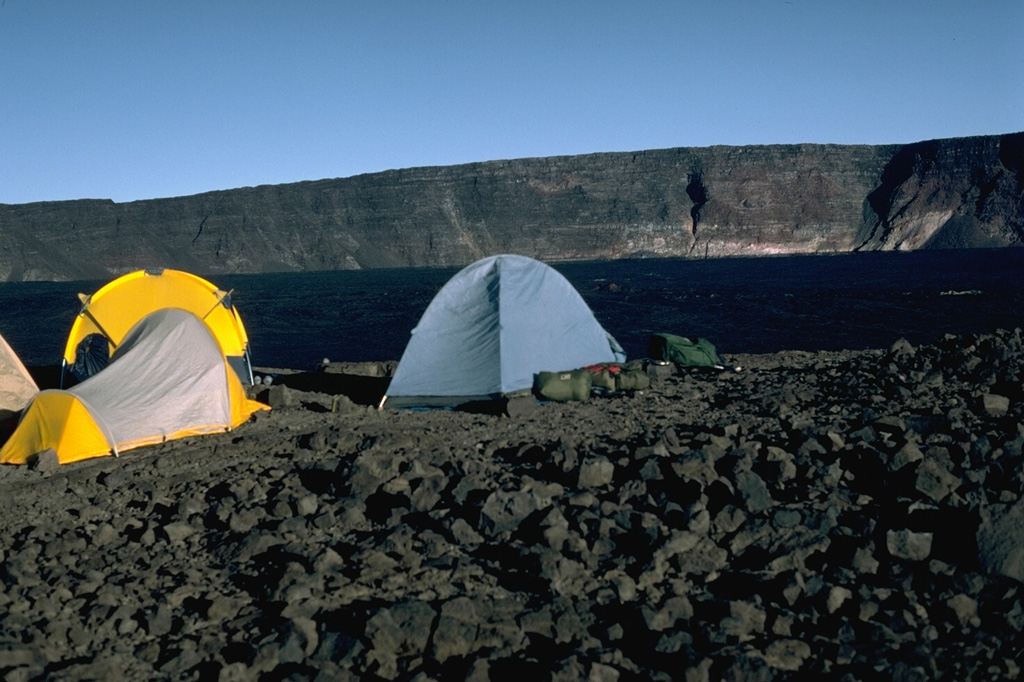 A campsite on the E rim of Mauna Loa's Moku’aweoweo caldera in 1987 has a view of the W rim that rises about 180 m above the lava flows from the 1984 eruption that covered the caldera floor. Lava flows from the 1975 eruption had also almost covered the entire floor. By this time, incremental filling of the caldera had almost halved the maximum recorded caldera depth of 320 m. Photo by Lee Siebert, 1987 (Smithsonian Institution).