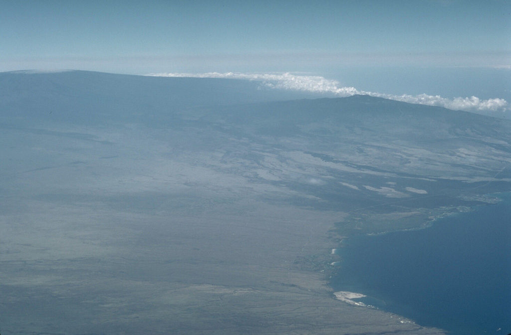 Hualalai volcano (upper right), the smallest of the young shield volcanoes on the island of Hawaii, rises above Kawaihae Bay in this aerial view from the north.  Hualalai shield volcano is perched on the western flank of the massive Mauna Loa shield volcano (upper left).  The latest eruption of Hualalai, during 1800-1801, produced lava flows from flank vents that reached the sea. Photo by Lee Siebert, 1987 (Smithsonian Institution).