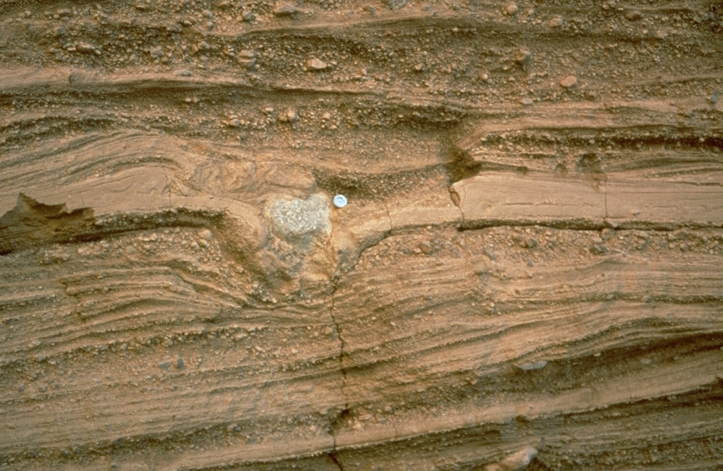 Pyroclastic surge deposits surround the Cerro Colorado maar of the Pinacate volcanic field in NW México. These thin beds (note the coin for scale) were formed by successive explosions that produced pyroclastic surges. The light-colored rock in the center of the photo is a ballistic block that impacted the surface of earlier surge deposits, compressing them and forming a small pit called a bomb sag. Photo by Richard Waitt, 1988 (U.S. Geological Survey).