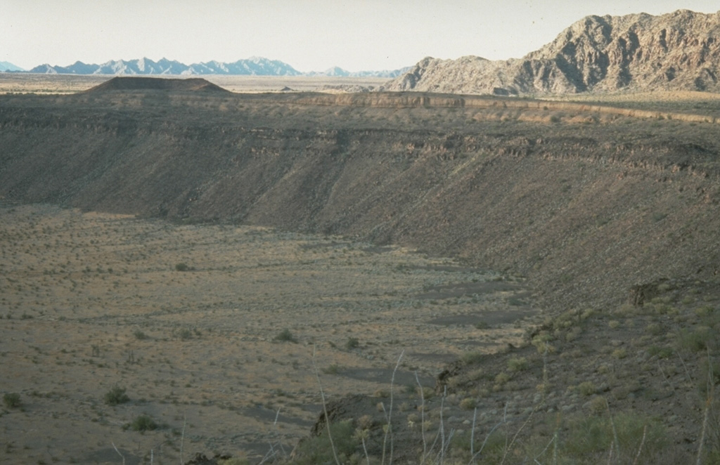 McDougal Crater, one of many Quaternary maars in the Pinacate volcanic field, formed within darker lava flow units near the top of the crater wall. Lighter-colored pyroclastic surge deposits form the crater rim and playa deposits formed on the crater floor. Crystalline rocks of the Sierrita el Temporal range are visible beyond the upper right crater rim to the north. Photo by Richard Waitt, 1988 (U.S. Geological Survey).