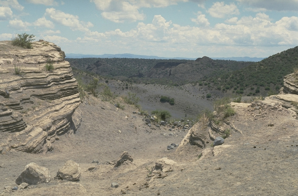 The La Breña-El Jagüey maar complex is one of the youngest features of the Durango volcanic field. This view looks SW from the NE rim of El Jagüey toward La Breña maar and shows the laminated pyroclastic surge beds emplaced during the maar-forming eruptions. El Jagüey and La Breña are two intersecting maars, 700 and 1,400 m wide, respectively.  Photo by Jim Luhr, 1988 (Smithsonian Institution).
