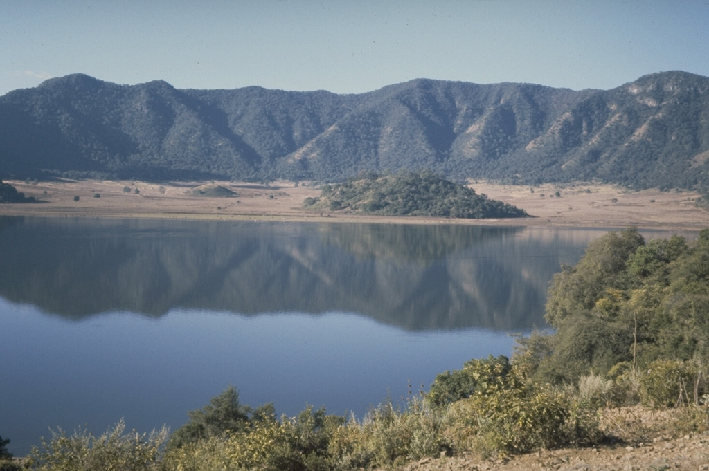 The oval-shaped, 5 x 2.5 km Tepetiltic caldera, seen here from the shore of a lake that occupies the older NE side of the caldera, was formed during two eruptive episodes that produced voluminous rhyodacitic airfall tephra and ashflows.  Formation of the compound caldera was closely followed by emplacement of two rhyodacitic lava domes on the eastern flank.  The low forested hill at the far side of the lake is a lava dome extruded near the intersection of the two calderas.  A second dome was emplaced to the left on the floor of the higher SW caldera. Photo by Jim Luhr, 1978 (Smithsonian Institution).