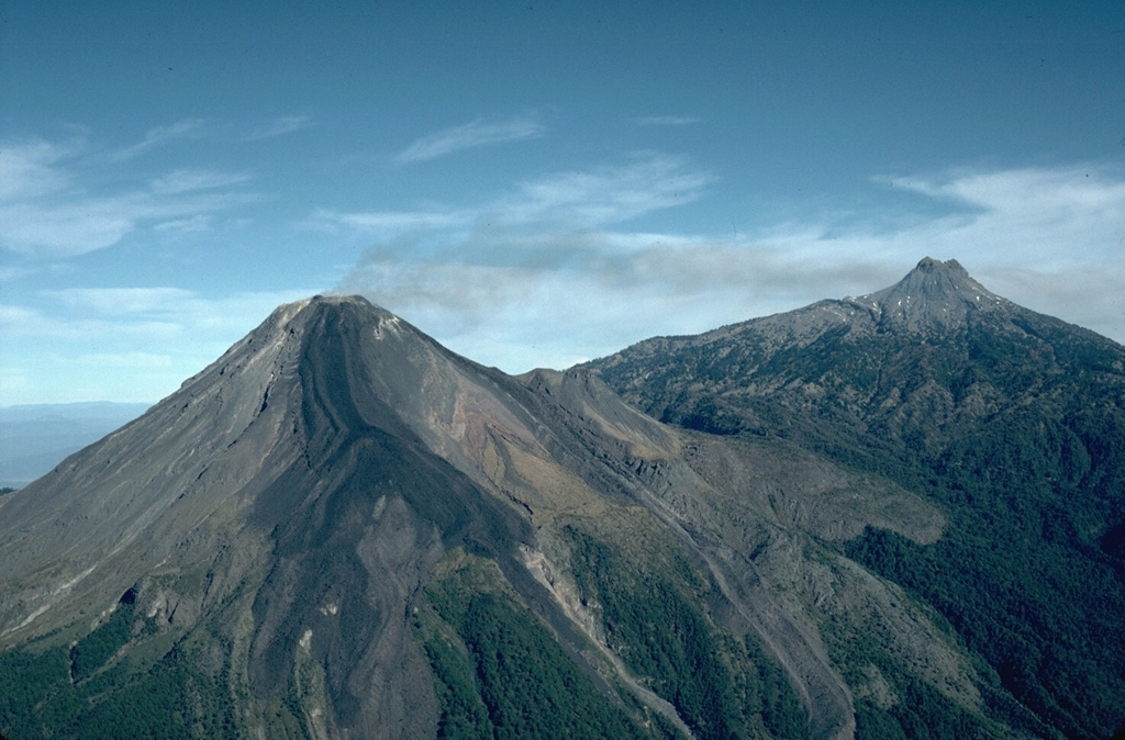 The Colima volcanic complex consists of the massive overlapping edifices Nevado de Colima (the highest point of the complex to the right) and Volcán de Colima (left). Volcán de Colima was constructed within a 5-km-wide caldera that opens to the south and has been the source of large debris avalanches. Frequent historical eruptions have included dome growth, explosive activity, pyroclastic flows, and lava flows. Photo by James Allan, 1981 (Smithsonian Institution).