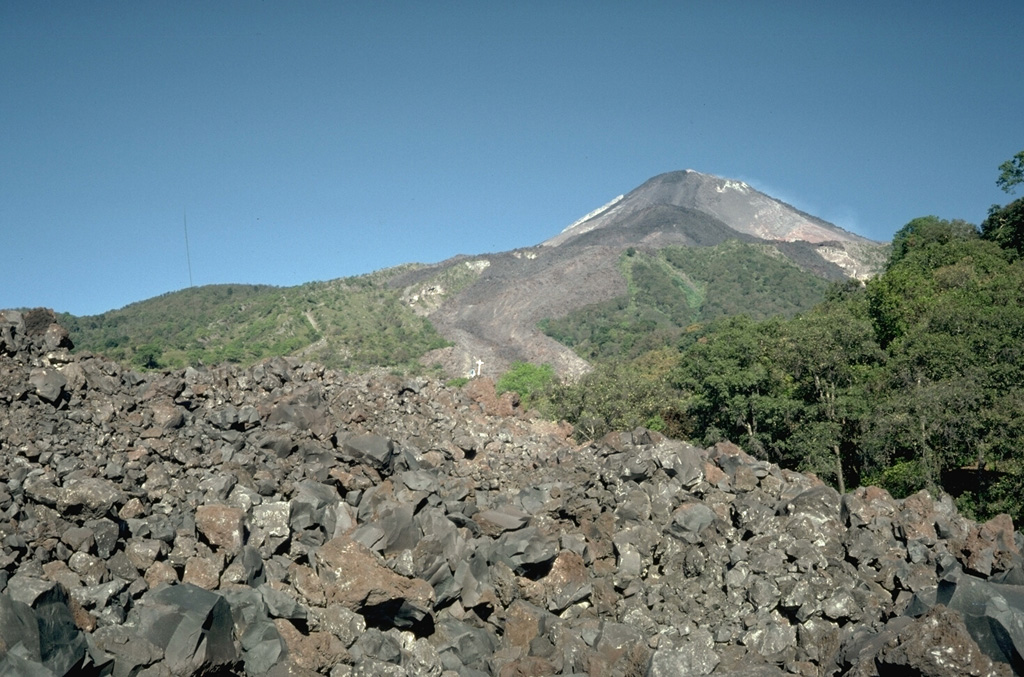 The lava flow in the foreground, which was emplaced during the 1975-76 eruption of Mexico's Colima volcano, can be seen descending through the vegetation at the center. The flow originated from the summit and traveled 3.5 km down the SE flank. The flow bifurcated on the upper slopes to form another lobe that traveled E, forming the darker area that descends into the vegetation at the upper right. Photo by Jim Luhr, 1983 (Smithsonian Institution).