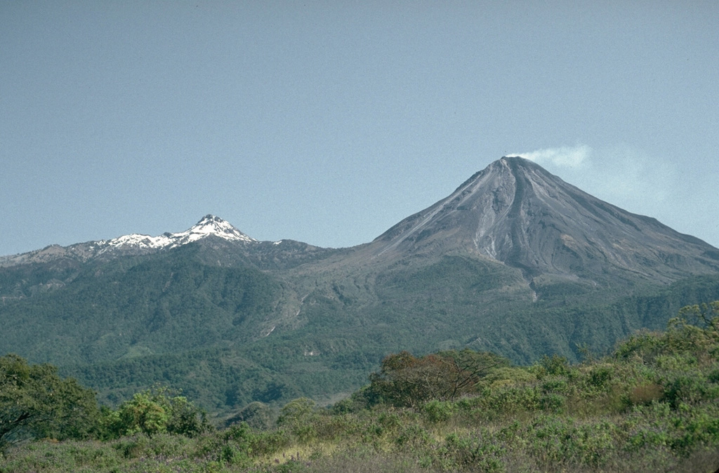 A weak plume from the summit of Mexico's Colima volcano in 1992 with snow-capped Nevado de Colima to the left; view from the WSW. Frequent eruptions have been recorded at Colima since the 16th century. Eruptions have been dominated during the past century by lava effusion associated with lava dome growth, explosive eruptions of varying magnitude, and frequent pyroclastic flows. Photo by Jim Luhr, 1992 (Smithsonian Institution).