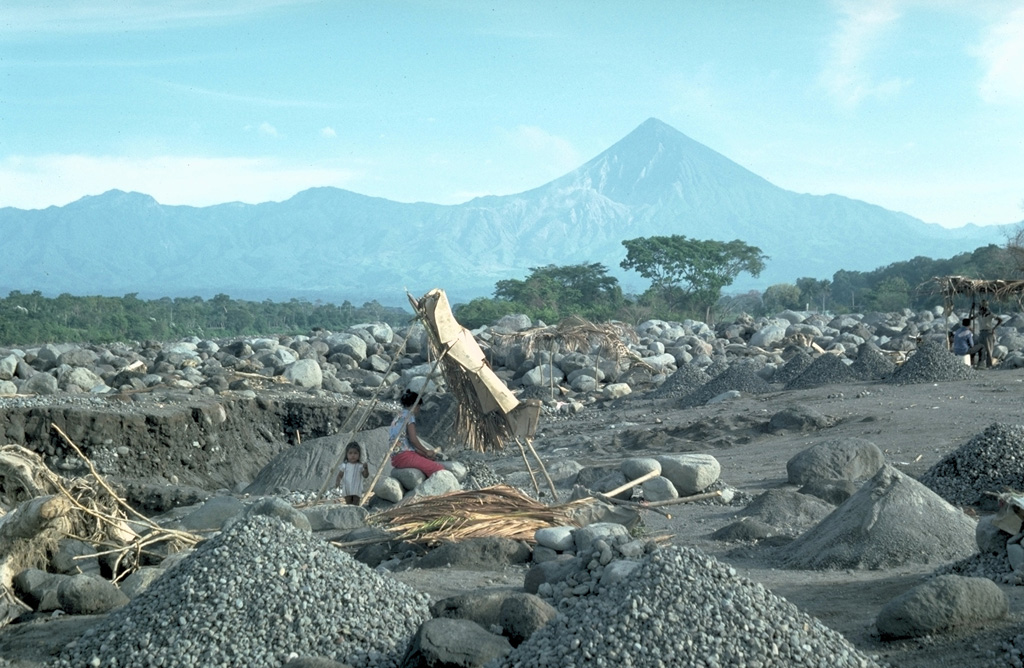 The debris from the Santiaguito lava dome (visible below and to the left of the summit of Santa María) severely impacts downslope river drainages. Frequent rainy season lahars descend the Río Samalá, seen here just south of the Pacific coastal highway. Major lahars on 28 August 1993 (three months prior to this photo) covered 200 m of the highway and filled the river channel to within 50 cm of the highway bridge. A small-scale construction aggregate operation can be seen in the foreground. Photo by Lee Siebert, 1993 (Smithsonian Institution).