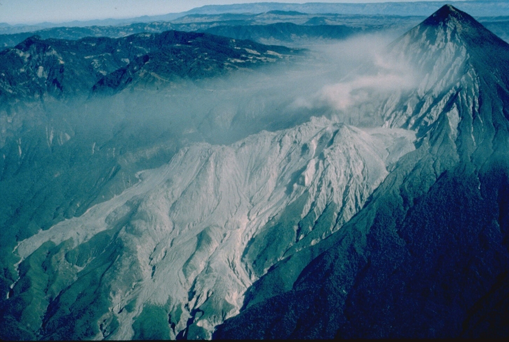 Two decades after a powerful explosive eruption in 1902 blasted a 1-km-wide crater through the SW flank of Santa María volcano, a lava dome began growing at the base of the crater.  Santiaguito lava dome, forming the elongated unvegetated ridge at the center of the photo, has been continually active since 1922.  Frequent explosive eruptions accompany episodic periods of more rapid lava dome growth and effusion of lava flows down the flanks of the dome.  The summit of Santiaguito is about 1300 m below that of Santa María at the upper right. Copyrighted photo by Katia and Maurice Krafft, 1983.