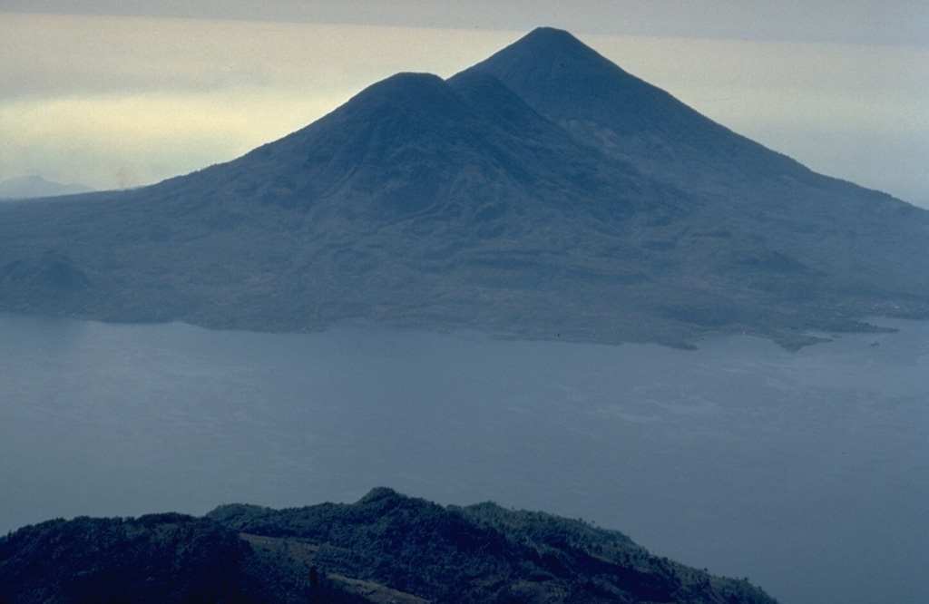 The twin volcanoes of Tolimán and Atitlán rise above the southern shores of Lake Atitlán in this view from the NNW.  The surface of Tolimán (left) is draped by prominent thick lava flows.  Many of the flows were erupted from vents on the volcano's flanks and form an irregular shoreline on the south side of Lake Atitlán.  A lava flow from the parasitic lava dome of Cerro de Oro on the north flank entered Lake Atitlán and is less than a few thousand years old.  No historical eruptions are known from Tolimán. Copyrighted photo by Katia and Maurice Krafft, 1983.
