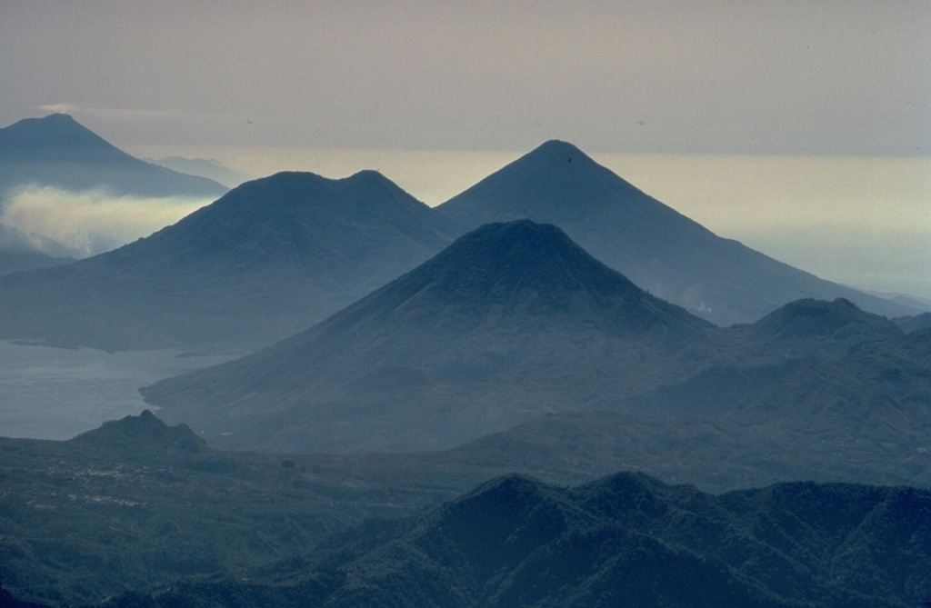 The twin volcanoes of Tolimán (left) and Atitlán (right) on the center skyline, along with San Pedro volcano in the foreground, rise above the southern shores of Lake Atitlán.  This aerial view looking along the chain of stratovolcanoes stretching across Guatemala, also shows Fuego volcano at the upper left.  The double-peaked Tolimán is somewhat older than the conical Atitlán volcano to its south. Copyrighted photo by Katia and Maurice Krafft, 1983.