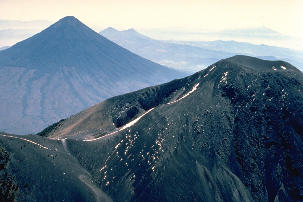 This photo looking SE across the volcanic chain of Guatemala shows Fuego volcano, lightly dusted with snow, in the foreground, and the symmetrical Agua volcano at the upper left.  Twin-peaked Pacaya volcano is located behind the right flank of Agua, and the broad massif in the distance above the summit of Fuego is Tecuamburro volcano.  Fuego and Pacaya are two of Guatemala's most active volcanoes. Copyrighted photo by Katia and Maurice Krafft, 1983.