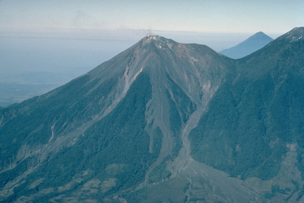 Volcán Fuego, one of Central America's most active volcanoes, is one of three large stratovolcanoes overlooking Guatemala's former capital, Antigua.  The scarp of an older edifice, Meseta, forms the shoulder between between Fuego and its twin volcano, Acatenango.  This view from the SE also shows Atitlán volcano above the saddle between Fuego and Acantenango.   Vigorous historical eruptions have been recorded at Fuego since 1524.  They have produced major ashfalls, along with occasional pyroclastic flows and lava flows.  Copyrighted photo by Katia and Maurice Krafft, 1983.