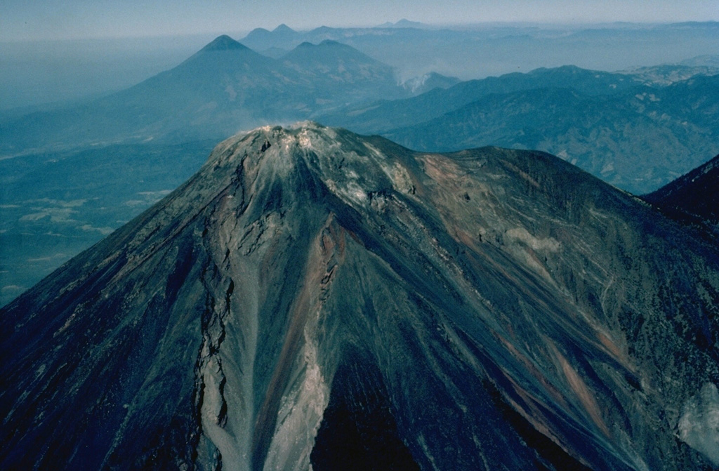 The summit of Fuego volcano is seen here from the ESE, looking along the chain of large stratovolcanoes stretching across Guatemala.  The shoulder to the right of Fuego is a remnant of Meseta volcano, whose growth and collapse preceded formation of the modern cone of Fuego.  Behind its summit are the paired volcanoes of conical Atitlán (left) and Tolimán, with a lower double summit.  Above the saddle between them are Pleistocene Santo Tomás (left) and the frequently active Santa María (right).  Tajumulco is on the skyline just right of center. Copyrighted photo by Katia and Maurice Krafft, 1983.