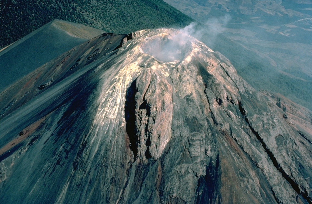 Steam rises above the summit of Volcán Fuego, one of Guatemala's most active volcanoes, seen here in March 1983 from the SW.  Thick masses of lava agglutinate and short lava flows armor parts of the upper cone.  The ash-covered shoulder left of the summit is the rim of Meseta.  The modern Fuego cone was constructed during the last 8500 years within an arcuate scarp left by collapse of Meseta volcano.  The forested slope at the upper left is that of Acatenango volcano. Copyrighted photo by Katia and Maurice Krafft, 1983.