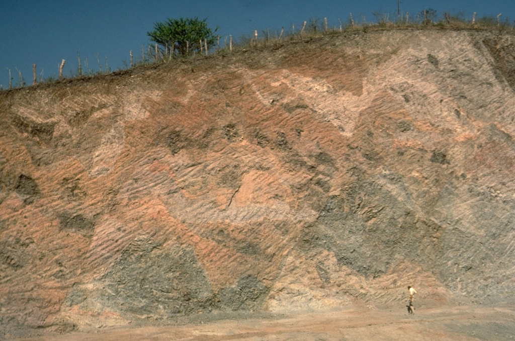 This roadcut provides a spectacular section through a debris avalanche deposit hummock of the Fuego-Acatenango complex in Guatemala. The pronounced color mottling is a common texture of debris-avalanche deposits and distinguishes them from deposits produced by other volcanic processes such as mudflows or pyroclastic flows. This texture results from the transport of coherent chunks of the volcano over long distances with only partial disaggregation. Individual lava flow blocks and pyroclastic units can be traced across small offsetting faults. Photo by Lee Siebert, 1988 (Smithsonian Institution).