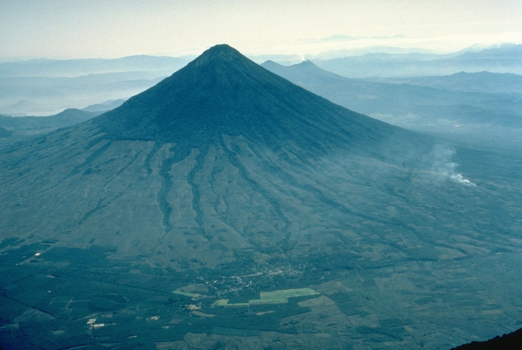 The symmetrical, forested Volcán de Agua stratovolcano forms a prominent backdrop to both the historic former capital city of Antigua Guatemala and Guatemala City, the present capital.  The 3760-m-high Agua volcano has a small, circular crater that is breached on the NNE side.  Agua has had no historical eruptions, but a devastating mudflow on September 11, 1541, destroyed the first Guatemalan capital city, now known as Ciudad Vieja.  This view from the NW also shows the twin-peaked Pacaya volcano behind the upper right flank of Agua. Copyrighted photo by Katia and Maurice Krafft, 1983.