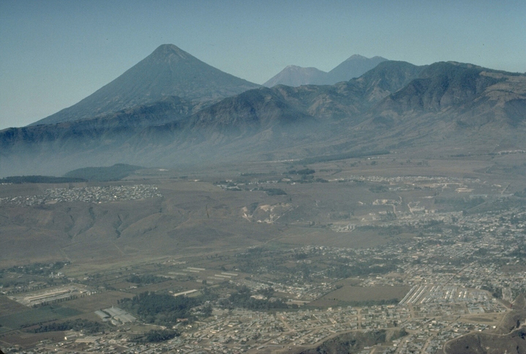 The beautifully symmetrical Agua volcano forms a prominent backdrop to Guatemala City in the foreground.  The twin volcanoes of Fuego (left) and Acatenango (right) appear to the right of Agua in this 1983 view from the NE.  No historical eruptions are known of Agua volcano, despite its youthful profile. Copyrighted photo by Katia and Maurice Krafft, 1983.