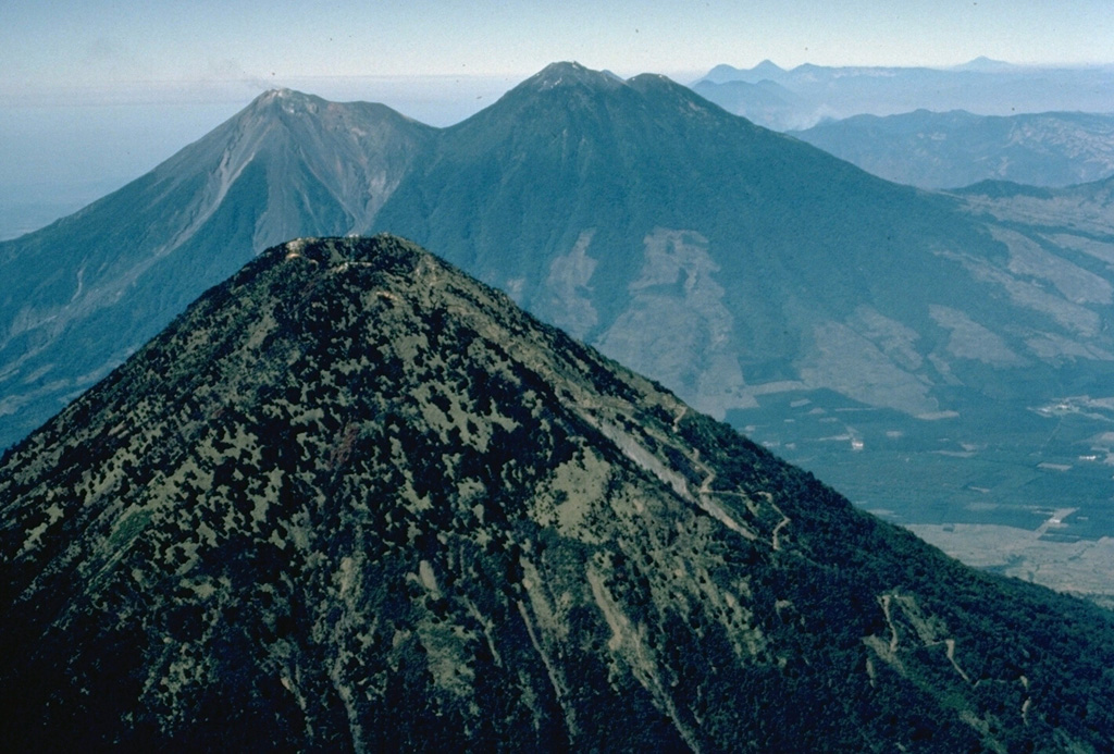 The forested summit of symmetrical Agua volcano appears in the foreground with the twin volcanoes of Fuego (left) and Acatenango (right) in the background.  The three stratovolcanoes overlook the historical city of Antigua Guatemala, whose outskirts appear on the right.  Agua has had no historical eruptions, in contrast to Acatenango and especially Fuego, which is one of the most active volcanoes of Guatemala.  This 1983 aerial view is from the west. Copyrighted photo by Katia and Maurice Krafft, 1983.