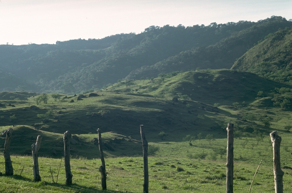 The hilly terrain in the foreground is part of a debris avalanche deposit that extends 25 km from the summit of Pacaya to the Pacific coastal plain. Part of the avalanche traveled more than 100 m up the side of the ridge in the background before coming to rest on the valley floor. The avalanche was accompanied by a pyroclastic surge whose deposits overlie the proximal 10 km.  Photo by Lee Siebert, 1988 (Smithsonian Institution).