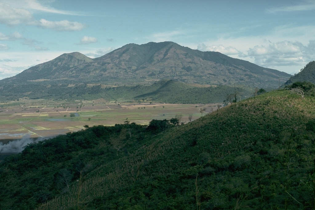 Suchitán volcano, seen here from the west on the rim of Retana caldera, is the highest of a group of closely spaced small volcanoes including scoria cones in SE Guatemala. The large peak to the left of the summit is Cerro Mataltepe; other scoria cones occur lower on the north flank. One of the more recent lava flows from Suchitán traveled through a low notch in the eastern caldera rim. The caldera once contained a lake, but now is used for agricultural land. Photo by Lee Siebert, 1993 (Smithsonian Institution).