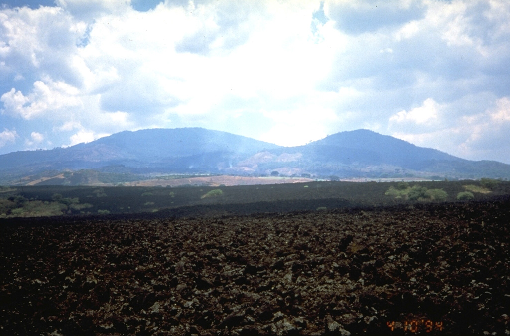The barren lava flow in the foreground was formed during a 1917 eruption from vents on the flank of Boquerón stratovolcano, the rounded peak at the left.  The lava flow cut the railway SW of Quezaltepeque.  The 1917 eruption also produced a small cinder cone in the summit crater of Boquerón.  Boquerón has grown within a 6-km-wide caldera whose western rim forms El Jabalí peak (right).  Three fracture zones that extend beyond the base of the volcano have been the locus for numerous flank eruptions of Santa Ana volcano.   Photo by Kristal Dorion, 1994 (U.S. Geological Survey).