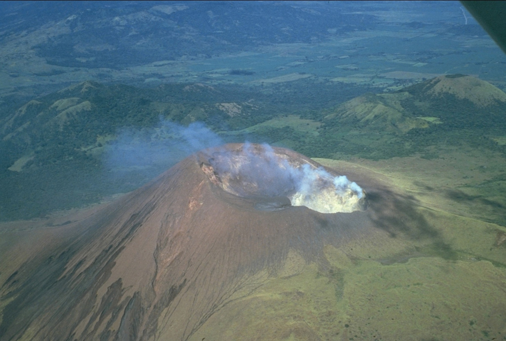 A prominent fumarole on the NE rim of the summit crater of Telica volcano produces a diffuse plume in this 1981 view.  The double summit crater is viewed from the SE with the grassy cone of Cerro de Aguero, the northernmost cone of the Telica volcanic complex, at the upper right.  The flanks of La Pelona caldera, part of the neighboring San Cristóbal volcanic complex, can be seen at the top of the photo across a low saddle. Photo by Jaime Incer, 1981.