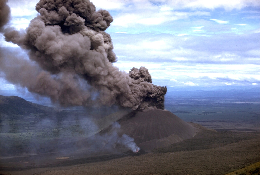 Winds deflect an ash column originating from the summit crater of Cerro Negro in 1968.  At the same time a vent on the lower south flank, which is emitting a thin, lighter-colored steam column in this view, fed a lava flow.  The advancing flow forms the black lobe extending to the lower left corner of the photo.  The 1968 eruption began on October 23 and continued until December 15. Photo by William Melson, 1968 (Smithsonian Institution).